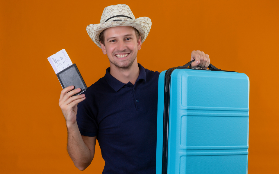 Um jovem e confiante com seu seguro viagem internacional, usando um chapéu de verão, está de pé com uma mala e segurando passagens aéreas. Ele olha diretamente para a câmera com um sorriso radiante e uma expressão feliz, enquanto o fundo laranja ressalta sua vibração positiva.